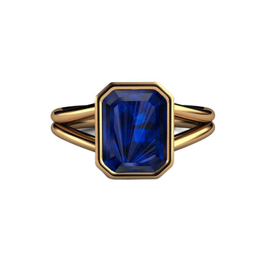 An emerald cut Blue Sapphire ring with a split shank double band and bezel set large lab grown blue sapphire in a yellow gold solitaire setting from Rare Earth Jewelry.