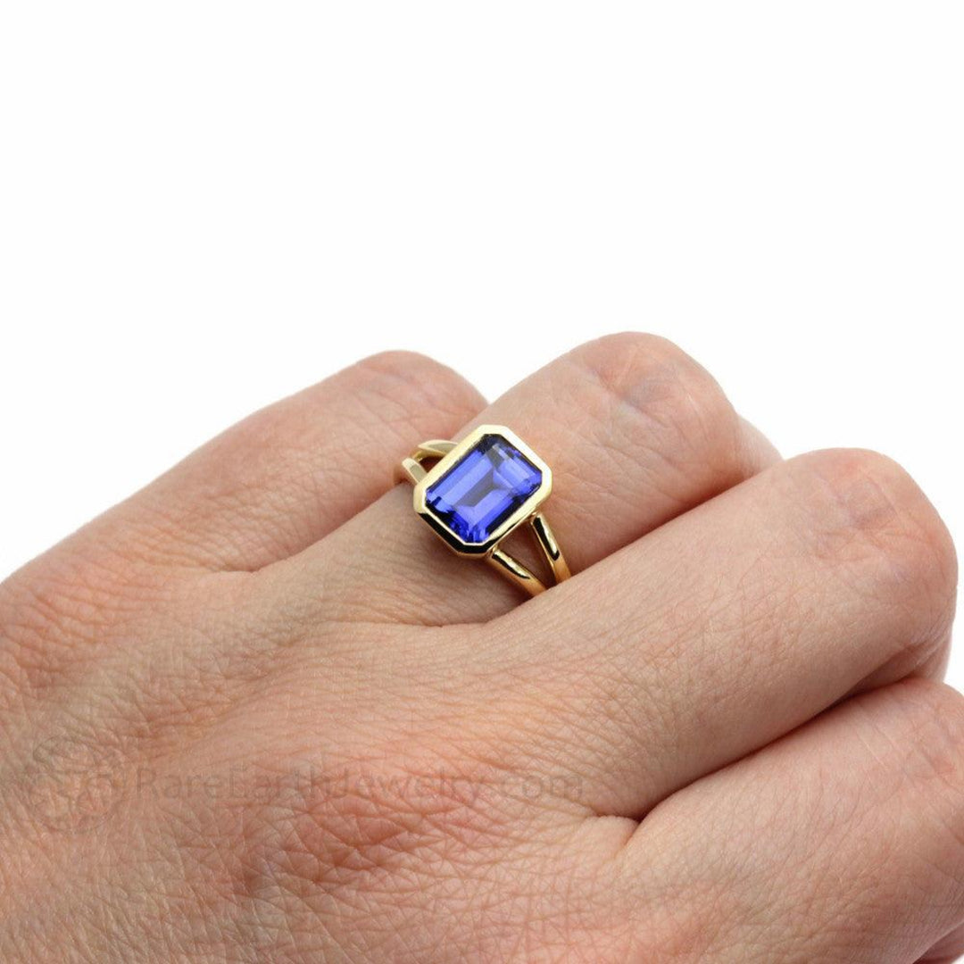 9x7mm emerald cut Blue sapphire engagement ring, bezel set solitaire with split shank in yellow gold ring on hand | Rare Earth Jewelry