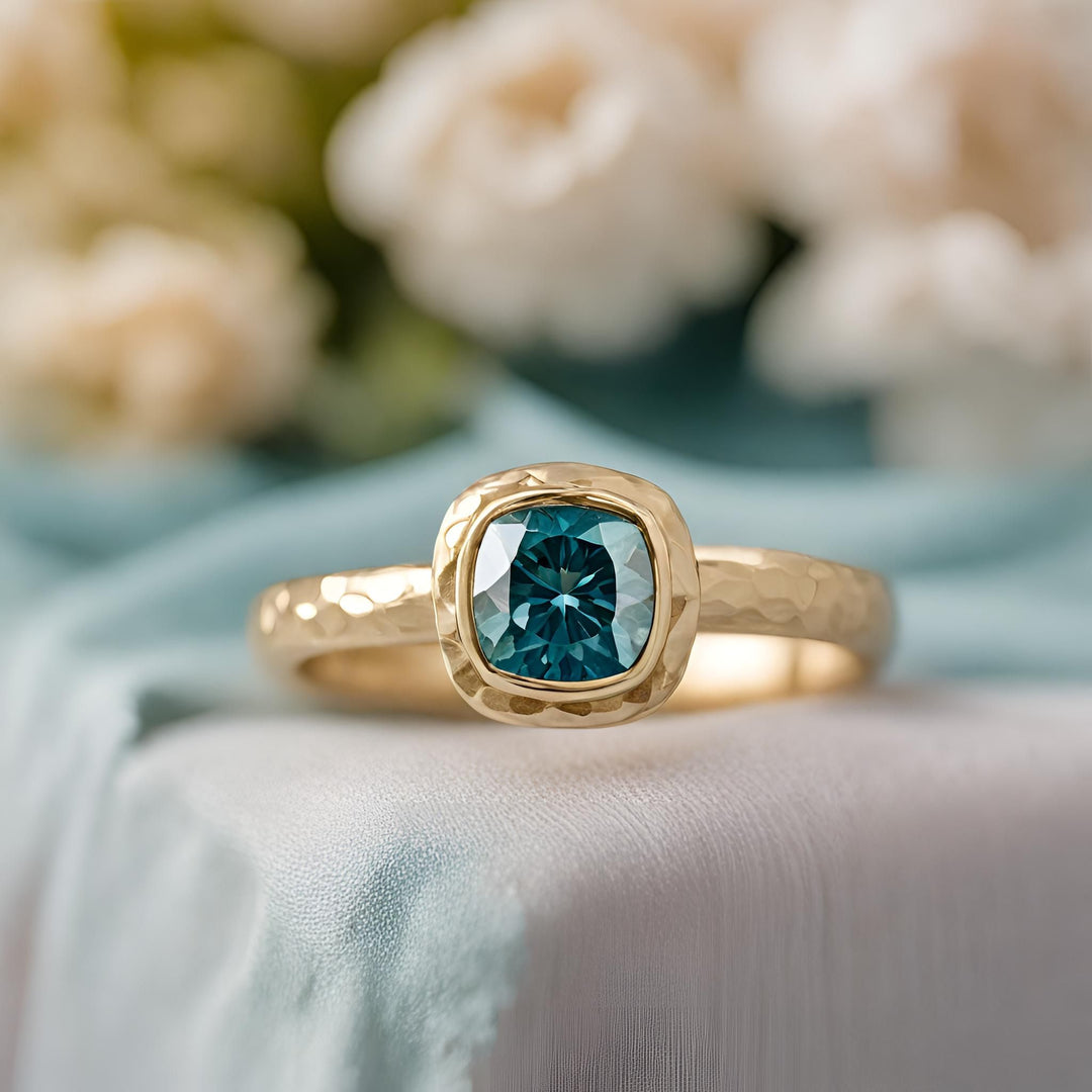 A cushion cut teal blue Moissanite engagement ring in a modern hammered bezel setting from Rare Earth Jewelry.