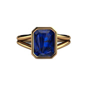 An emerald cut Blue Sapphire ring with a split shank band and bezel set large lab grown blue sapphire in a yellow gold solitaire setting..