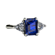 An emerald cut lab grown blue sapphire engagement ring in a three stone style with white sapphire trillions in gold and platinum from Rare Earth Jewelry.