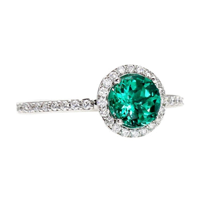 A round lab created emerald ring in a diamond halo engagement ring style with diamonds in a halo and on the band in gold or platinum.  May birthstone ring from Rare Earth Jewelry.