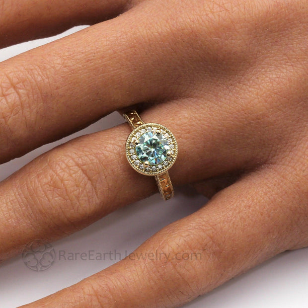 A pastel Blue Moissanite Engagement Ring in a vintage style diamond halo design with engraved band shown on the hand photo from Rare Earth Jewelry
